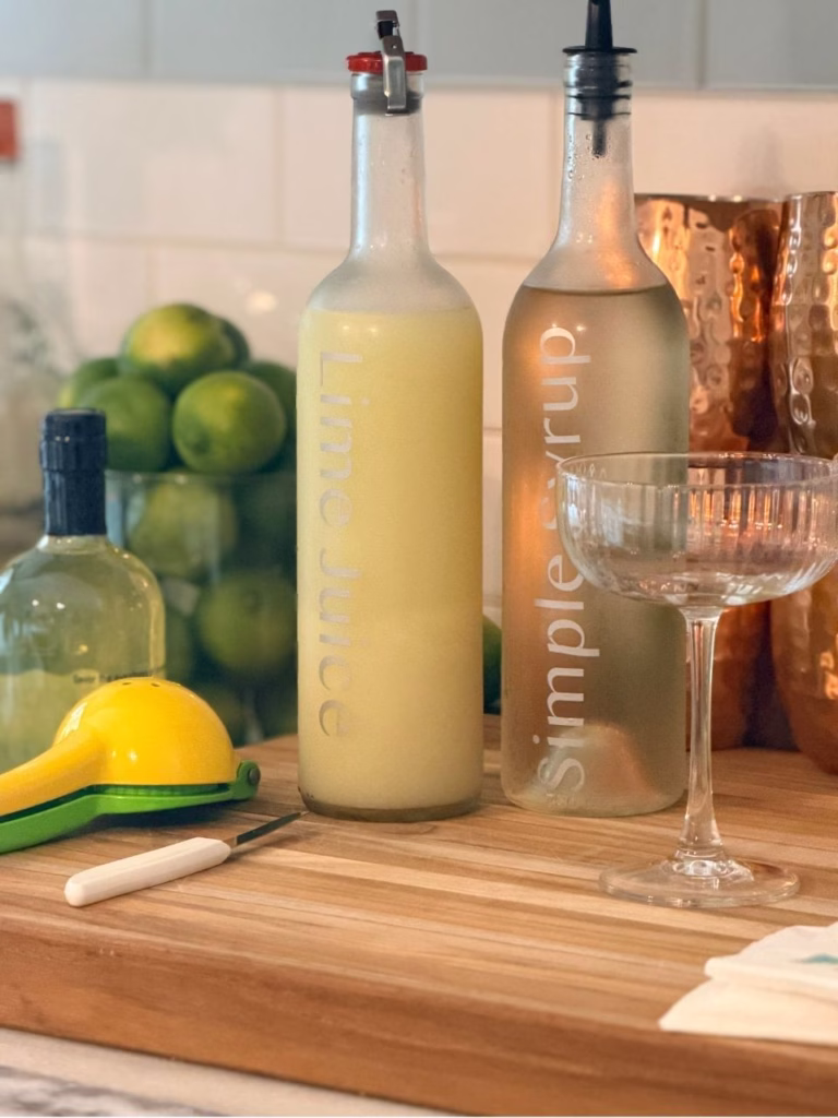 A cutting board with two bottles on it, limes in the background, and a juicer to the side